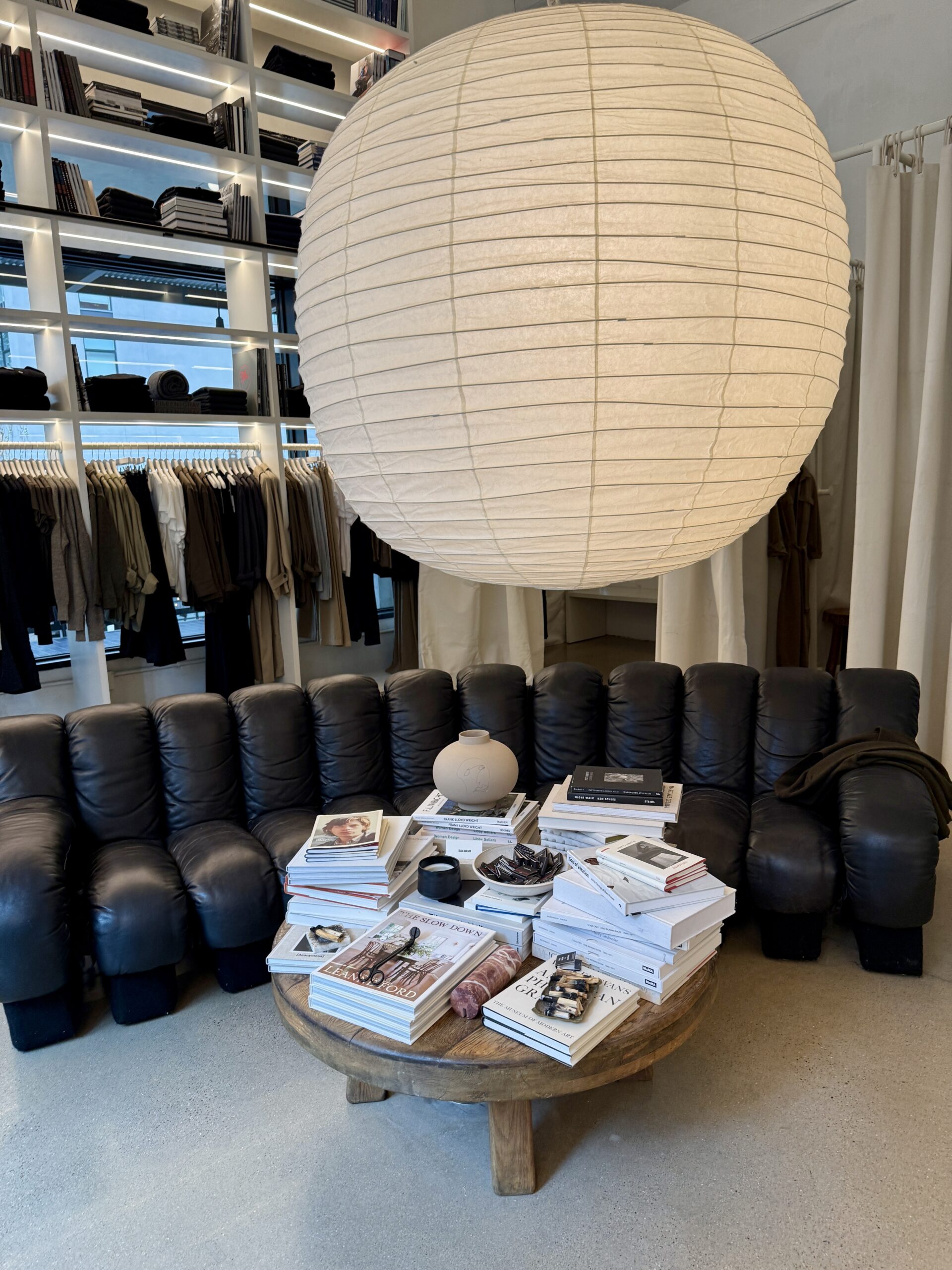 Interior of a minimalist boutique or studio with a large white paper lantern hanging over a black leather modular sofa. A round wooden coffee table in front of the sofa is stacked with neatly arranged art and design books, magazines, and small objects. Behind the seating area, built-in shelves display folded clothing and books, with neutral-toned garments hanging on racks, creating a calm, curated, editorial atmosphere.